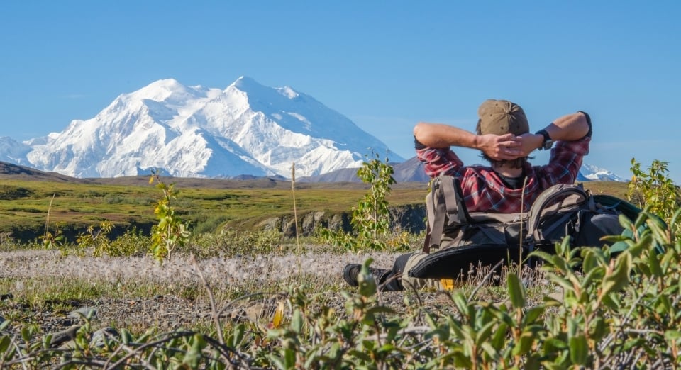 Man lying on his back against his backpack while looking up at the snow-covered mountain at Denali National Park