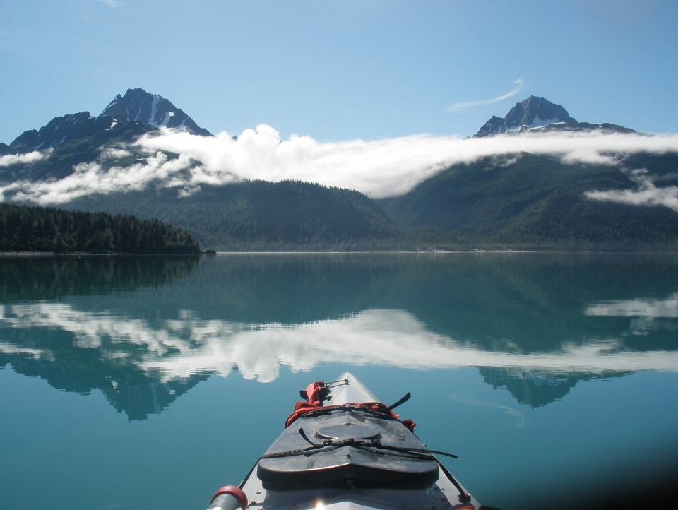 Kayak on still water. Ahead is a range of snow-capped mountains, with clouds wafting around them