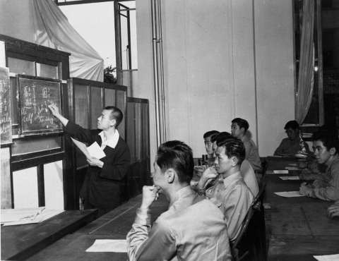 Black and white photo of a class at MISLS. An instructor writes on a blackboard in front of two rows of students sitting at tables.