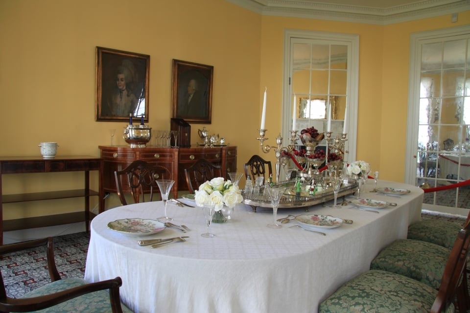 A formal dining room table is set with a vase of white flowers and long white taper candles. On the pale yellow walls hang two portraits.