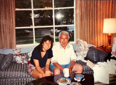 A young woman sits on a couch with her grandfather, both smiling at the camera.