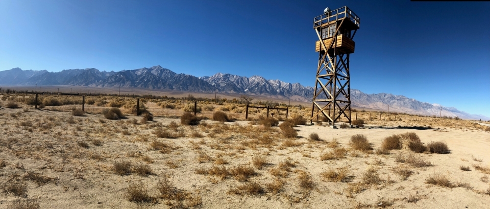 A wooden guard tower looms over a desolate, sandy landscape. A mountain range hangs in the background