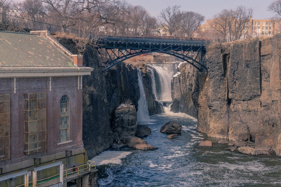 A waterfall cascades down a stone cliff. A brick building is nestled next to the base of the falls, and bridge looms above