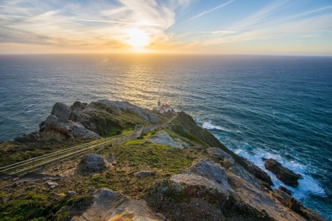 Four white-sided, red-roofed structures sit on a rocky headland above the Pacific Ocean at the base of a long stairway. Above and to the left of the lighthouse, the sun filters through wispy clouds as it descends toward the horizon