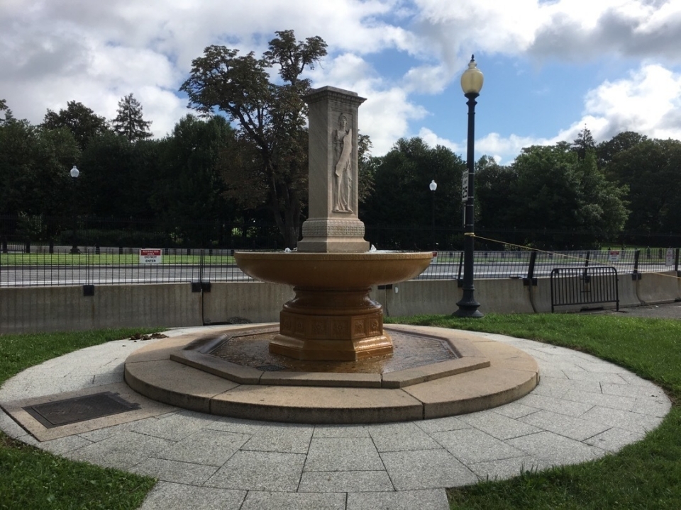A circular fountain with a rectangular stone sculpture depicting Francis Davis Millet in the middle