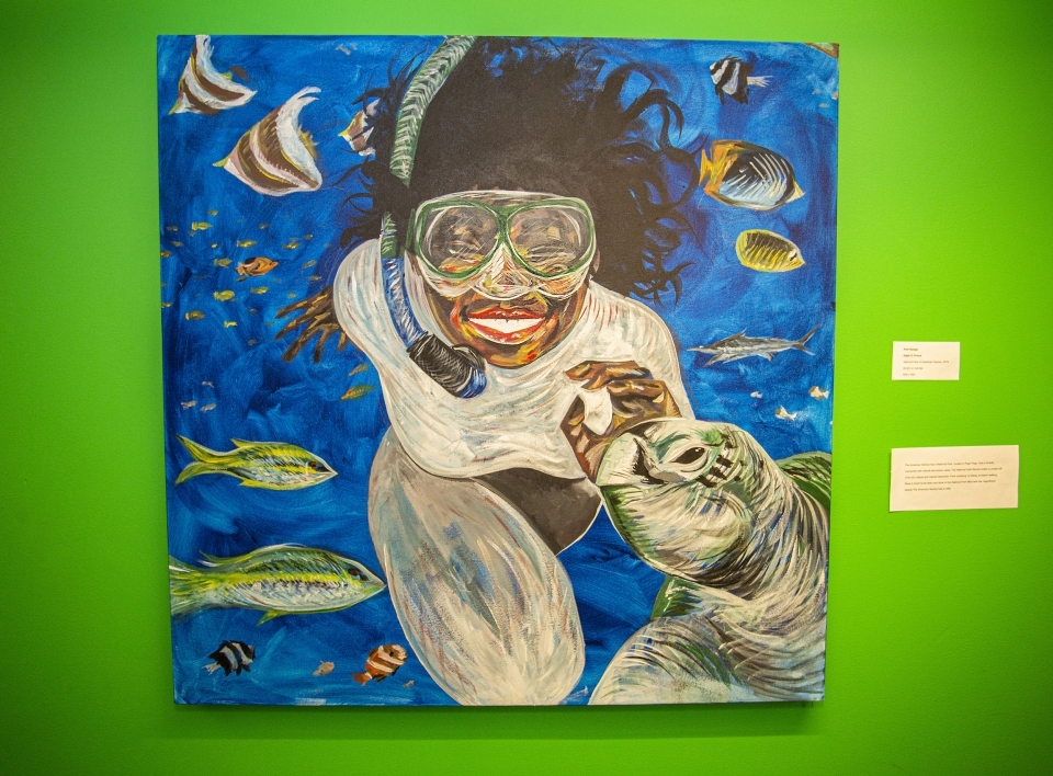 Painting against a bright green wall, depicting a woman snorkeling and smiling. A variety of fish swim around her, and a sea turtle seems to smile in the bottom right corner.