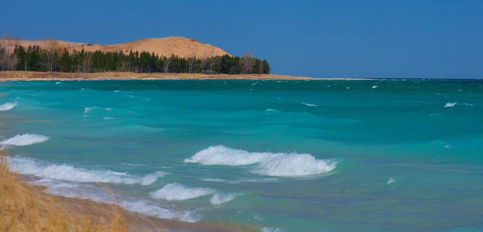 Bright blue waters along a warm brown shoreline. In the background, a large dune crests over the water