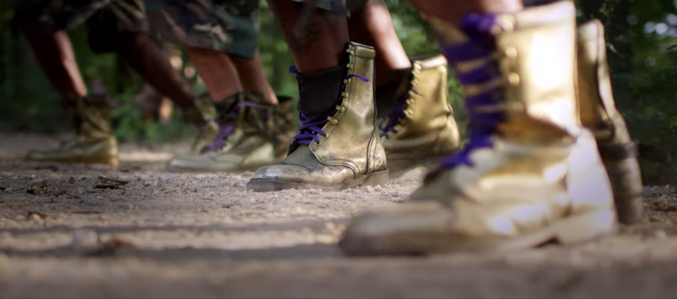 A row of gilded combat boots stomp on a dirt ground