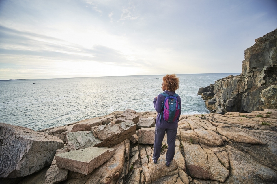 A woman stands on a rocks overlooking water. She is wearing a rain proof athletic jacket and a backpack.