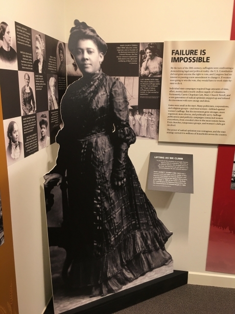 Full body cut out of Mary Burnett Talbert with a long, black dress and bun hair style. The cut out is next to the Lifting as We Climb panel.